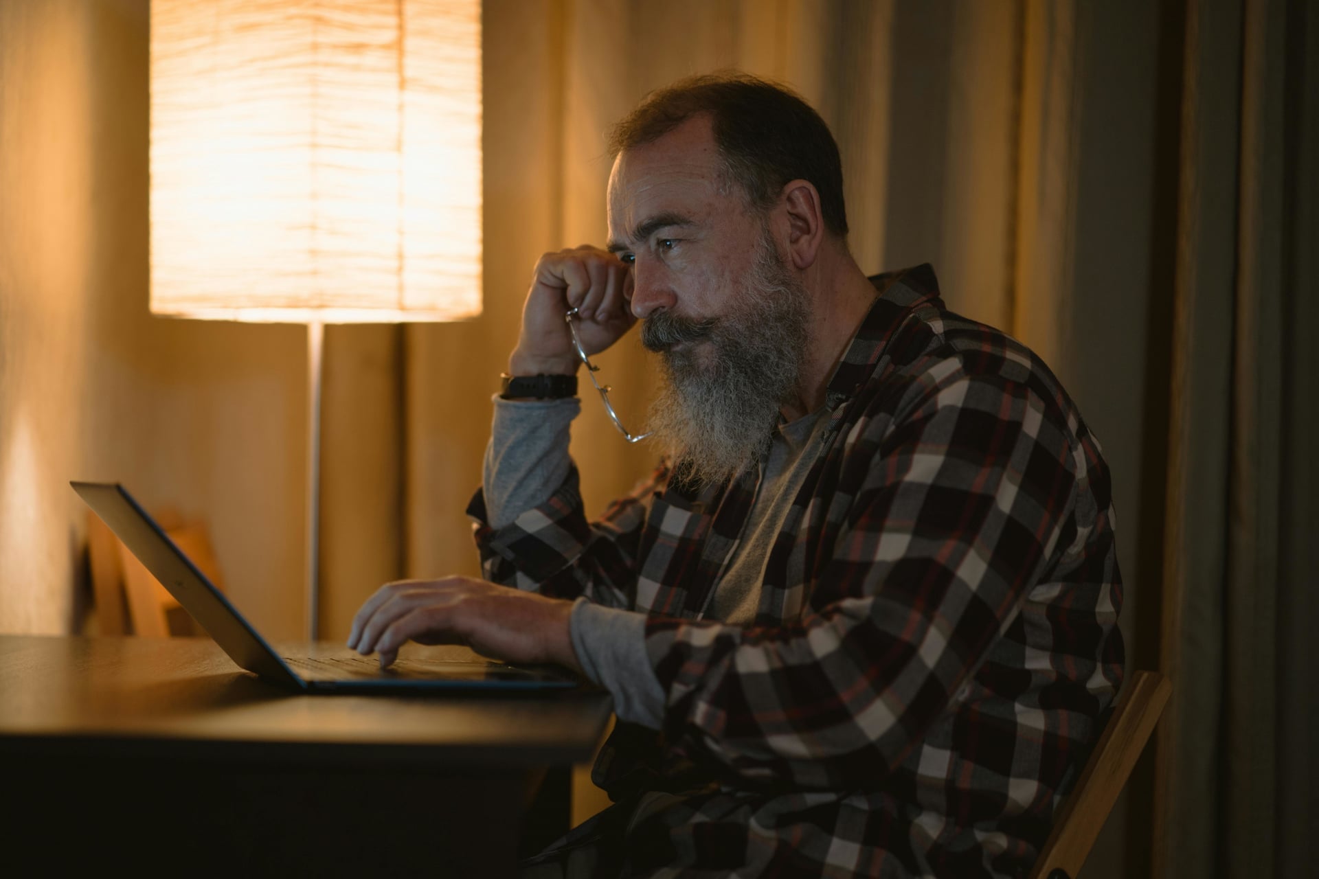Mann med skjegg sitter ettertenksomt ved laptop i varmt lampelys